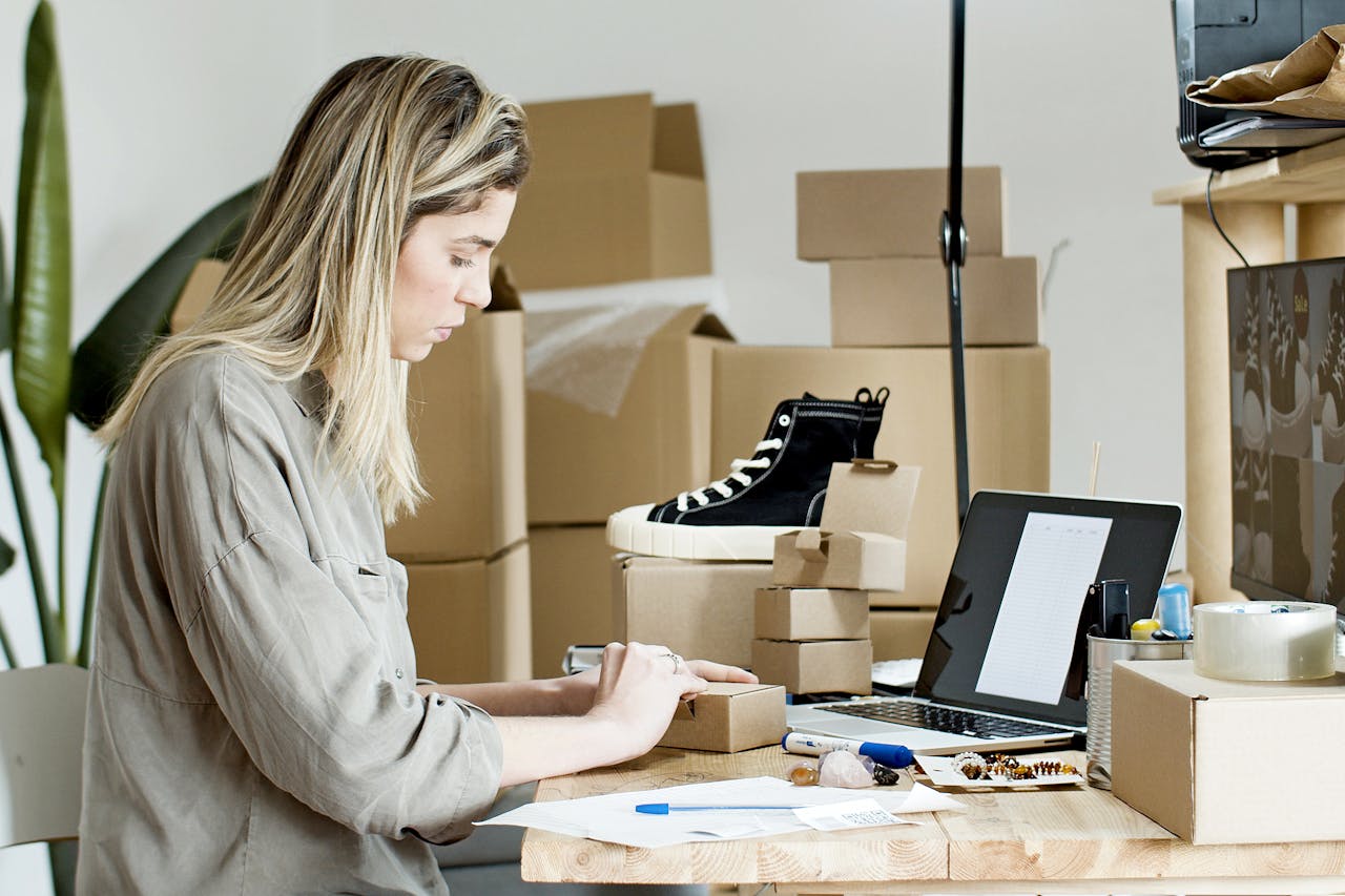 about-us-02 A woman packing boxes for her online store, surrounded by packaging materials and a laptop.